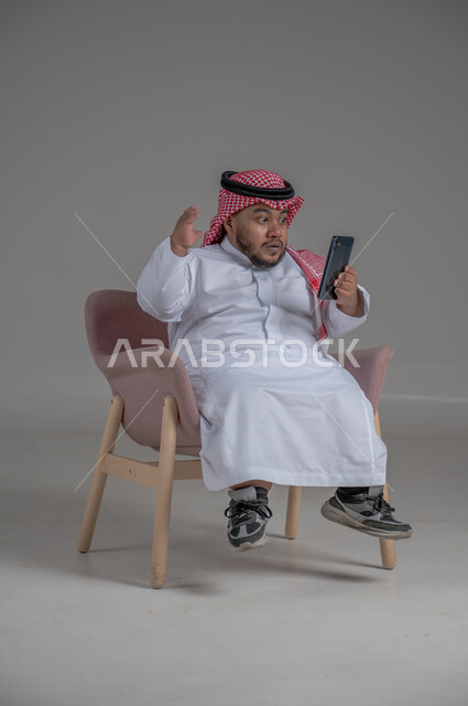 قضاء وقت ممتع, بورتريه لرجل عربي خليجي سعودي قصير القامة يجلس على كرسي مريح ويستخدم الهاتف المحمول ، استخدام البرامج والتطبيقات الالكترونية، التسوق الالكتروني عبر الانترنت، تصفح المواقع الالكترونية والتواصل الاجتماعي، خلفية رمادية