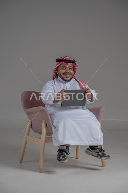 مفهوم إدارة المشاريع عن بعد, إستخدام اجهزة تقنية حديثة، بورتريه لرجل عربي خليجي سعودي قصير القامة يرتدي الشماغ والثوب التقليدي يجلس على الكرسي يقوم بانجاز مهام العمل عبر جهاز التابلت, خلفية رمادية
