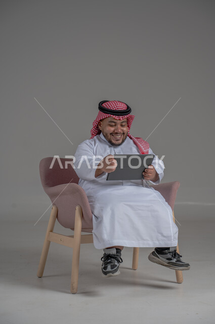 Remote project management concept, using modern technology devices, portrait of a short Saudi Arabian Gulf man wearing a traditional keffiyeh and thobe sitting on a chair completing work tasks via a tablet, gray background