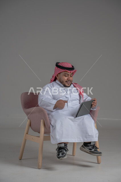 Remote project management concept, using modern technology devices, portrait of a short Saudi Arabian Gulf man wearing a traditional keffiyeh and thobe sitting on a chair completing work tasks via a tablet, gray background