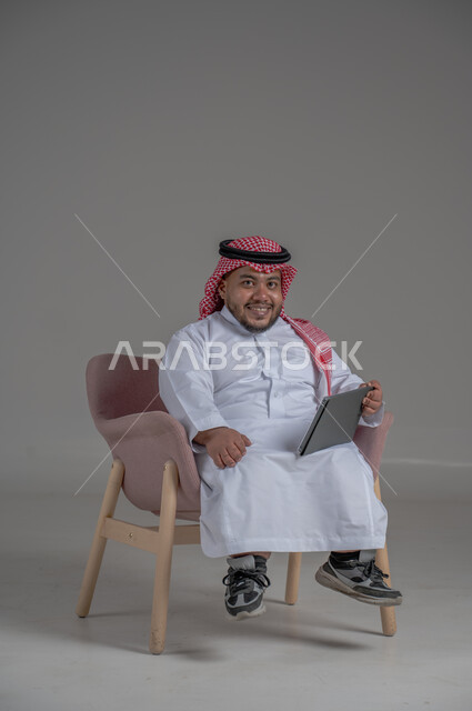 Remote project management concept, using modern technology devices, portrait of a short Saudi Arabian Gulf man wearing a traditional keffiyeh and thobe sitting on a chair completing work tasks via a tablet, gray background