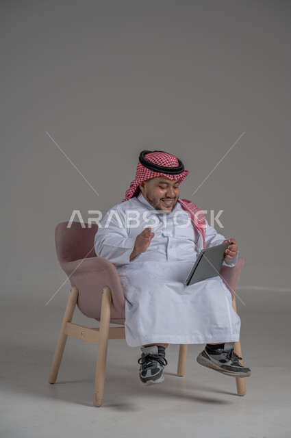 مفهوم إدارة المشاريع عن بعد, إستخدام اجهزة تقنية حديثة، بورتريه لرجل عربي خليجي سعودي قصير القامة يرتدي الشماغ والثوب التقليدي يجلس على الكرسي يقوم بانجاز مهام العمل عبر جهاز التابلت, خلفية رمادية