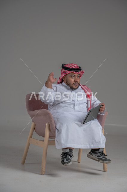 Remote project management concept, using modern technology devices, portrait of a short Saudi Arabian Gulf man wearing a traditional keffiyeh and thobe sitting on a chair completing work tasks via a tablet, gray background