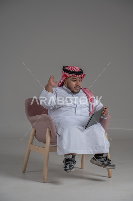 Remote project management concept, using modern technology devices, portrait of a short Saudi Arabian Gulf man wearing a traditional keffiyeh and thobe sitting on a chair completing work tasks via a tablet, gray background