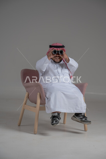 الشغف وحب الاستكشاف والمغامرة، البحث عن شيء ما باستخدام الناظور، مفهوم رؤية الأشياء البعيدة، بورتريه لرجل عربي خليجي سعودي قصير القامة يرتدي الثوب التقليدي والشماغ يجلس على كرسي مريح ينظر من خلال التلسكوب, حلم تحقيق الأهداف والطموحات المستقبلية, خلفية رمادية