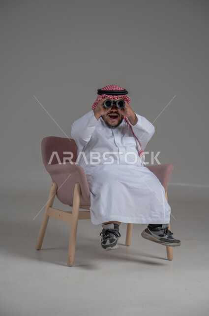الشغف وحب الاستكشاف والمغامرة، البحث عن شيء ما باستخدام الناظور، مفهوم رؤية الأشياء البعيدة، بورتريه لرجل عربي خليجي سعودي قصير القامة يرتدي الثوب التقليدي والشماغ يجلس على كرسي مريح ينظر من خلال التلسكوب, حلم تحقيق الأهداف والطموحات المستقبلية, خلفية رمادية