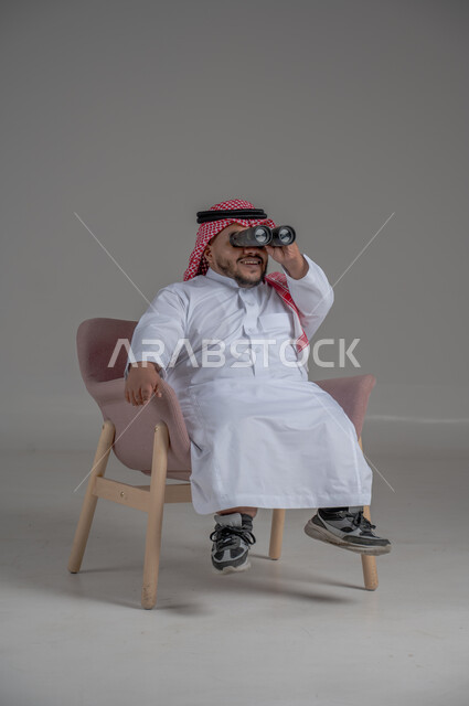 الشغف وحب الاستكشاف والمغامرة، البحث عن شيء ما باستخدام الناظور، مفهوم رؤية الأشياء البعيدة، بورتريه لرجل عربي خليجي سعودي قصير القامة يرتدي الثوب التقليدي والشماغ يجلس على كرسي مريح ينظر من خلال التلسكوب, حلم تحقيق الأهداف والطموحات المستقبلية, خلفية رمادية