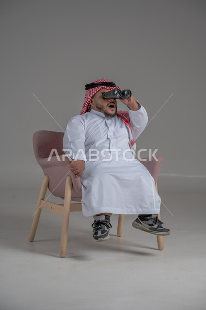 الشغف وحب الاستكشاف والمغامرة، البحث عن شيء ما باستخدام الناظور، مفهوم رؤية الأشياء البعيدة، بورتريه لرجل عربي خليجي سعودي قصير القامة يرتدي الثوب التقليدي والشماغ يجلس على كرسي مريح ينظر من خلال التلسكوب, حلم تحقيق الأهداف والطموحات المستقبلية, خلفية رمادية
