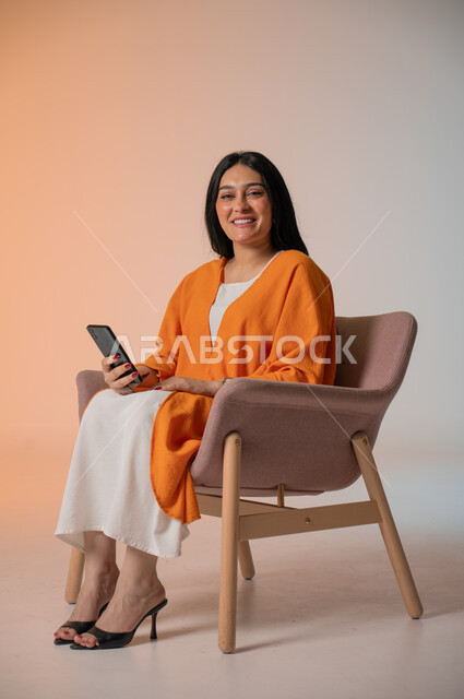 دمج التكنولوجيا المتطورة في الحياة اليومية، إجراء مكالمات الفيديو والتواصل مع الأهل والأصدقاء، بورتريه لامرأة عربية خليجية سعودية ترتدي الزي الكاجوال تجلس على كرسي وتحمل بيدها الهاتف المحمول وتنظر إلى الكاميرا بإيماءات السرور، التقاط صور السيلفي، خلفية ملونة