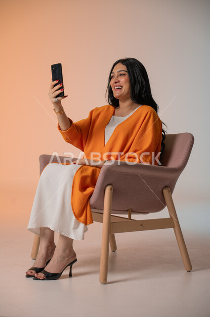 دمج التكنولوجيا المتطورة في الحياة اليومية، إجراء مكالمات الفيديو والتواصل مع الأهل والأصدقاء، بورتريه لامرأة أنيقة عربية خليجية سعودية ترتدي الزي الكاجوال تجلس على كرسي وتحمل بيدها الهاتف المحمول وتنظر إليه بإيماءات السرور، التقاط صور السيلفي، خلفية ملونة