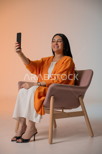 دمج التكنولوجيا المتطورة في الحياة اليومية، إجراء مكالمات الفيديو والتواصل مع الأهل والأصدقاء، بورتريه لامرأة أنيقة عربية خليجية سعودية ترتدي الزي الكاجوال تجلس على كرسي وتحمل بيدها الهاتف المحمول وتنظر إليه بإيماءات السرور، التقاط صور السيلفي، خلفية ملونة