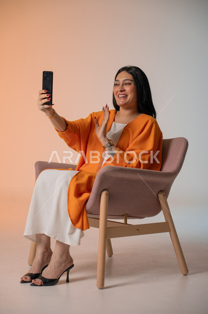 دمج التكنولوجيا المتطورة في الحياة اليومية، إجراء مكالمات الفيديو والتواصل مع الأهل والأصدقاء، بورتريه لامرأة أنيقة عربية خليجية سعودية ترتدي الزي الكاجوال تجلس على كرسي وتحمل بيدها الهاتف المحمول وتنظر إليه بإيماءات السرور، التقاط صور السيلفي، خلفية ملونة