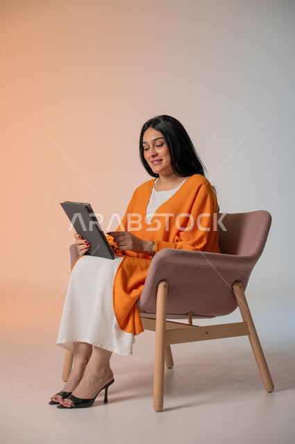 إجراء مكالمات الفيديو والتواصل مع الأهل والأصدقاء، متابعة آخر المستجدات، دمج التكنولوجيا المتطورة في الحياة اليومية، بورتريه لامرأة عربية خليجية سعودية ترتدي الزي الكاجوال تجلس على كرسي وتحمل بيدها جهاز التابلت وتنظر إليه بإيماءات السرور والاندماج، خلفية ملونة