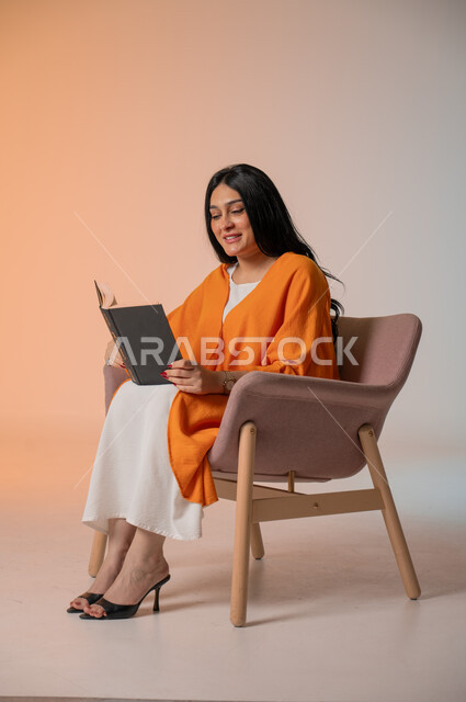 تنمية شغف المطالعة والثقافة، قراءة الكتب المتنوعة وتزويد الذخيرة الفكرية، بورتريه لامرأة أنيقة عربية خليجية سعودية ترتدي الزي الكاجوال تجلس على كرسي مريح وتحمل بيدها كتاب وتطالع فيه بإيماءات الاندماج والتركيز، أهمية تثقيف الأنثى لبناء مجمتع ناجح، خلفية ملونة