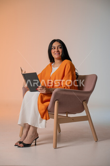 تنمية شغف المطالعة والثقافة، قراءة الكتب المتنوعة وتزويد الذخيرة الفكرية، بورتريه لامرأة أنيقة عربية خليجية سعودية ترتدي الزي الكاجوال تجلس على كرسي مريح وتحمل بيدها كتاب وتطالع فيه بإيماءات الاندماج والتركيز، أهمية تثقيف الأنثى لبناء مجمتع ناجح، خلفية ملونة