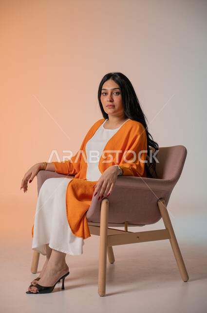 مفهوم الأناقة والاعتناء بالمظهر الخارجي، بورتريه لامرأة أنيقة عربية خليجية سعودية ترتدي الزي الكاجوال تجلس على كرسي مريح وتنظر إلى الكاميرا بإيماءات الثقة بالنفس والبشاشة، الاهتمام بالفاشن ومواكبة آخر صيحات الموضة، أزياء نسائية عصرية خليجية، خلفية ملونة