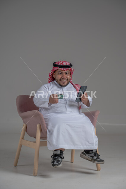 استخدام تطبيقات الجوال للدفع والشراء، التقنيات الحديثة المتطورة، تسديد الفواتير بالبطاقة الإلكترونية، بورتريه لرجل عربي خليجي سعودي قصير القامة يرتدي الشماغ والثوب التقليدي يجلس على كرسي مريح و يمسك بيده الهاتف المحمول وبطاقة الإئتمان البنكية، خلفية رمادية