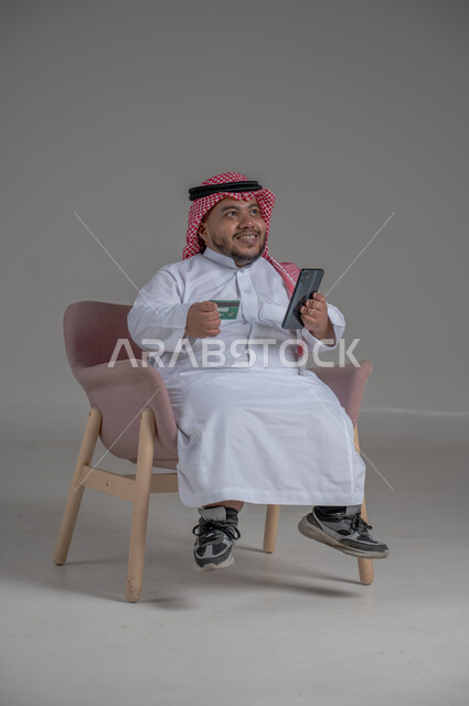 Using mobile applications for payment and purchase, advanced modern technologies, paying bills with electronic card, portrait of a short Saudi Arabian Gulf man wearing a traditional shemagh and thobe sitting on a comfortable chair and holding a mobile phone and a bank credit card in his hand, gray background