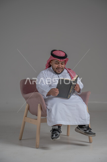 قضاء وقت الفراغ بالتعلم والثقافة، حب المطالعة و الاستكشاف ,القراءة بتركيز وتمعن، مفهوم محو الامية، بورتريه لرجل عربي خليجي سعودي قصير القامة يرتدي الثوب التقليدي و الشماغ يجلس على كرسي مريح يحمل بيده كتاب علمي تثقيفي, خلفية رمادية