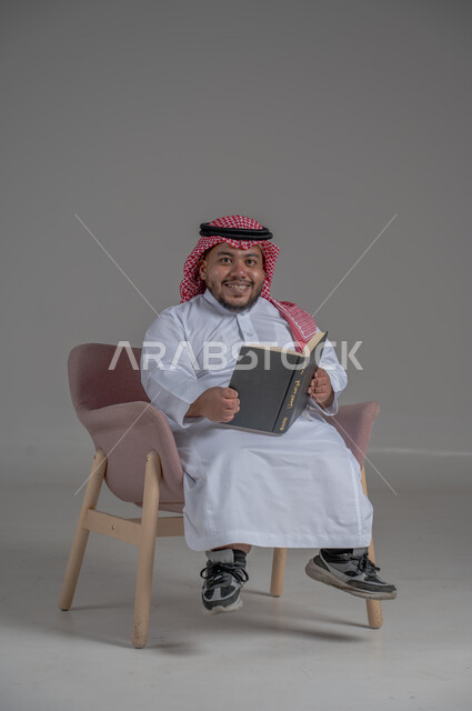 قضاء وقت الفراغ بالتعلم والثقافة، حب المطالعة و الاستكشاف ,القراءة بتركيز وتمعن، مفهوم محو الامية، بورتريه لرجل عربي خليجي سعودي قصير القامة يرتدي الثوب التقليدي و الشماغ يجلس على كرسي مريح يحمل بيده كتاب علمي تثقيفي, خلفية رمادية