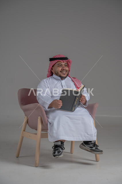 قضاء وقت الفراغ بالتعلم والثقافة، حب المطالعة و الاستكشاف ,القراءة بتركيز وتمعن، مفهوم محو الامية، بورتريه لرجل عربي خليجي سعودي قصير القامة يرتدي الثوب التقليدي و الشماغ يجلس على كرسي مريح يحمل بيده كتاب علمي تثقيفي, خلفية رمادية