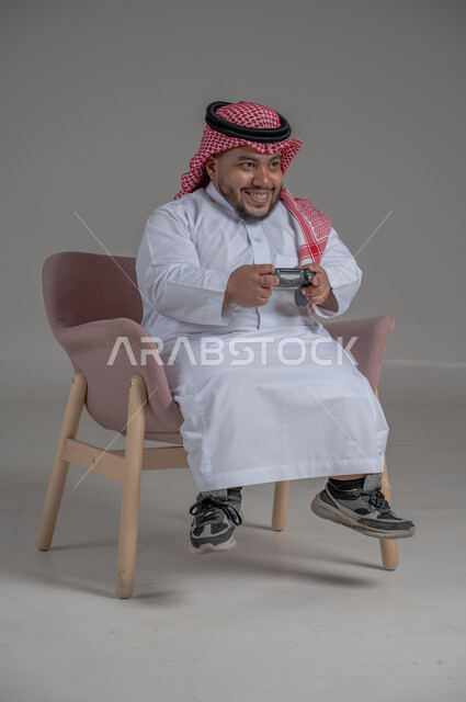 تقنيات حديثة مسلية, قضاء وقت ممتع ومسلي، التفاعل والاندماج مع ألعاب البلايستيشن، بورتريه لرجل عربي سعودي خليجي قصير القامة يرتدي الثوب التقليدي والشماغ يجلس على كرسي مريح ويحمل بيده عصا التحكم ويلعب بألعاب الفيديو، خلفية رمادية
