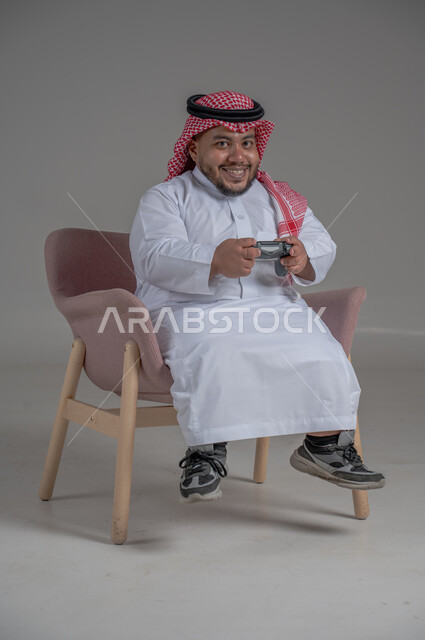 تقنيات حديثة مسلية, قضاء وقت ممتع ومسلي، التفاعل والاندماج مع ألعاب البلايستيشن، بورتريه لرجل عربي سعودي خليجي قصير القامة يرتدي الثوب التقليدي والشماغ يجلس على كرسي مريح ويحمل بيده عصا التحكم ويلعب بألعاب الفيديو، خلفية رمادية