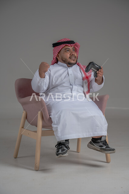 تقنيات حديثة مسلية, قضاء وقت ممتع ومسلي، التفاعل والاندماج مع ألعاب البلايستيشن، بورتريه لرجل عربي سعودي خليجي قصير القامة يرتدي الثوب التقليدي والشماغ يجلس على كرسي مريح ويحمل بيده عصا التحكم ويلعب بألعاب الفيديو، خلفية رمادية
