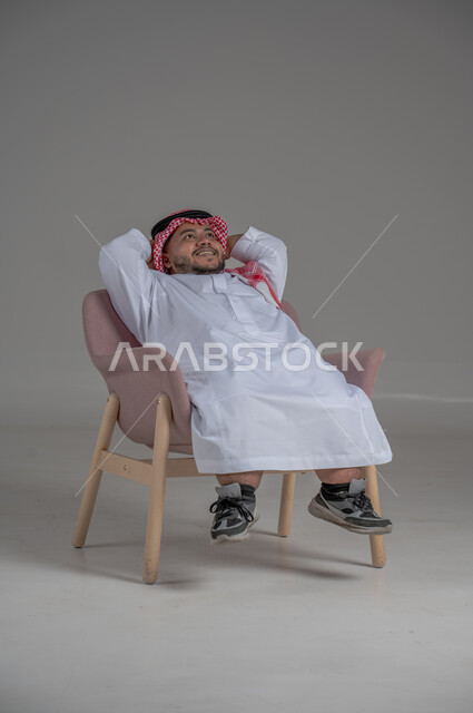 الشعور بالراحة والطمأنينة، مفهوم الاسترخاء والاستمتاع بالأجواء الهادئة، بورتريه لرجل عربي خليجي سعودي قصير القامة يرتدي الثوب التقليدي و الشماغ يجلس على الكرسي ويضع يديه فوق رأسه، خلفية رمادية