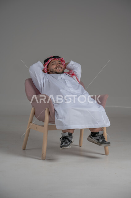 الشعور بالراحة والطمأنينة، مفهوم الاسترخاء والاستمتاع بالأجواء الهادئة، بورتريه لرجل عربي خليجي سعودي قصير القامة يرتدي الثوب التقليدي و الشماغ يجلس على الكرسي ويضع يديه فوق رأسه، خلفية رمادية