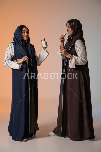 العروض الإعلانية التسويقية لبراندات الميكاب والعطور، الاهتمام بالفاشن ومواكبة الموضة، أزياء نسائية عصرية للمحجبات، بورتريه لشابتين عربيتين خليجيتين سعوديتين محجبتين ترتديان العباءات الملونة تمسكان مستحضرات التجميل وزجاجة العطر، الجسم بالطول الكامل، خلفية ملونة