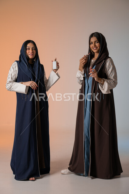 العروض الإعلانية التسويقية لبراندات الميكاب والعطور، الاهتمام بالفاشن ومواكبة الموضة، أزياء نسائية عصرية للمحجبات، بورتريه لشابتين عربيتين خليجيتين سعوديتين محجبتين ترتديان العباءات الملونة تمسكان مستحضرات التجميل وزجاجة العطر، الجسم بالطول الكامل، خلفية ملونة