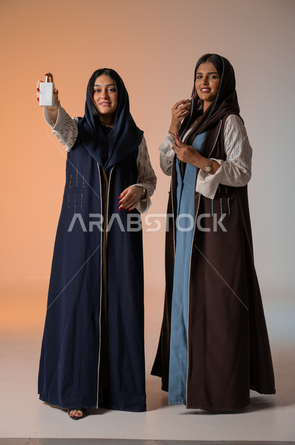العروض الإعلانية التسويقية لبراندات الميكاب والعطور، الاهتمام بالفاشن ومواكبة الموضة، أزياء نسائية عصرية للمحجبات، بورتريه لشابتين عربيتين خليجيتين سعوديتين محجبتين ترتديان العباءات الملونة تمسكان مستحضرات التجميل وزجاجة العطر، الجسم بالطول الكامل، خلفية ملونة