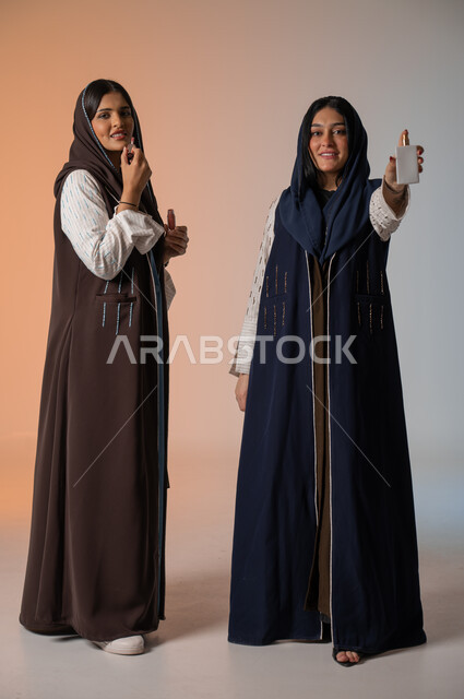 العروض الإعلانية التسويقية لبراندات الميكاب والعطور، الاهتمام بالفاشن ومواكبة الموضة، أزياء نسائية عصرية للمحجبات، بورتريه لشابتين عربيتين خليجيتين سعوديتين محجبتين ترتديان العباءات الملونة تمسكان مستحضرات التجميل وزجاجة العطر، الجسم بالطول الكامل، خلفية ملونة