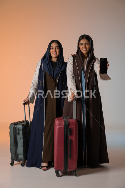 عرض شاشة هاتف محمول سوداء فارغة، بورتريه مقرب لصديقتين عربيتين خليجيتين سعوديتين محجبتين ترتديان العباءات الملونة تقومان بالاستعداد للانطلاق في رحلة ممتعة بإيماءات الحماس، تجهيز حقائب الأمتعة والاستمتاع  بالسفر رفقة الأصدقاء، الجسم بالطول الكامل، خلفية ملونة