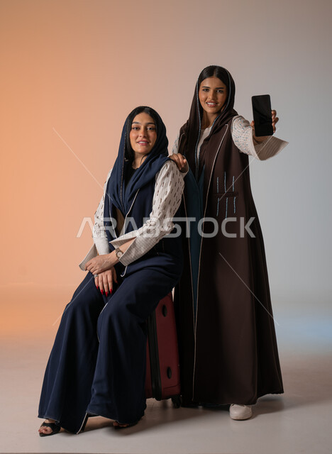 عرض شاشة هاتف محمول سوداء فارغة، بورتريه مقرب لصديقتين عربيتين خليجيتين سعوديتين محجبتين ترتديان العباءات الملونة تقومان بالاستعداد للانطلاق في رحلة ممتعة بإيماءات الحماس، تجهيز حقائب الأمتعة والاستمتاع  بالسفر رفقة الأصدقاء، الجسم بالطول الكامل، خلفية ملونة