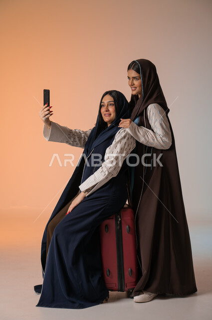 تجهيز حقيبة الأمتعة والاستعداد للسفر رفقة الأصدقاء، الاهتمام بالفاشن ومواكبة الموضة، أزياء نسائية عصرية للمحجبات، بورتريه لشابتين عربيتين خليجيتين سعوديتين محجبتين ترتديان العباءات الملونة تقومان بالتقاط صور السيلفي عبر الجوال، الجسم بالطول الكامل، خلفية ملونة