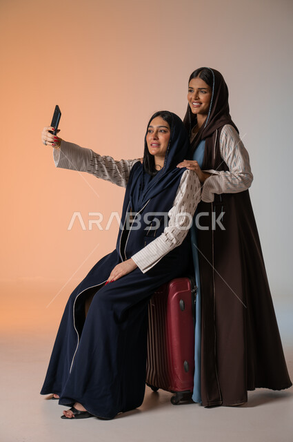 تجهيز حقيبة الأمتعة والاستعداد للسفر رفقة الأصدقاء، الاهتمام بالفاشن ومواكبة الموضة، أزياء نسائية عصرية للمحجبات، بورتريه لشابتين عربيتين خليجيتين سعوديتين محجبتين ترتديان العباءات الملونة تقومان بالتقاط صور السيلفي عبر الجوال، الجسم بالطول الكامل، خلفية ملونة