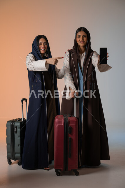  الإشارة إلى الهاتف المحمول وعرض شاشة سوداء فارغة، بورتريه مقرب لصديقتين عربيتين خليجيتين سعوديتين محجبتين ترتديان العباءات الملونة تقومان بالاستعداد للانطلاق في رحلة ممتعة، تجهيز حقائب الأمتعة ومفهوم السفر رفقة الأصدقاء، الجسم بالطول الكامل، خلفية ملونة