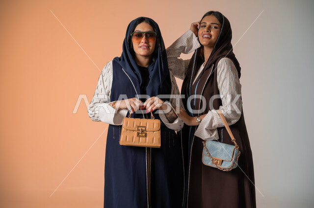 اقتناء اكسسوارات ومنتجات لبراندات عالمية، أزياء نسائية عصرية للمحجبات، الاهتمام بالفاشن ومواكبة آخر صيحات الموضة، بورتريه مقرب لصديقتين عربيتين خليجيتين سعوديتين محجبتين ترتديان العباءات الملونة وتمسكان الحقائب الجلدية الفاخرة، وضع النظارات الشمسية، خلفية ملونة