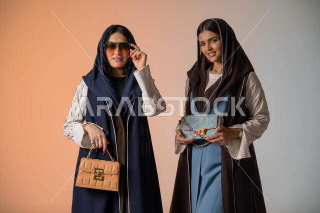 اقتناء اكسسوارات ومنتجات لبراندات عالمية، أزياء نسائية عصرية للمحجبات، الاهتمام بالفاشن ومواكبة آخر صيحات الموضة، بورتريه مقرب لصديقتين عربيتين خليجيتين سعوديتين محجبتين ترتديان العباءات الملونة وتمسكان الحقائب الجلدية الفاخرة، وضع النظارات الشمسية، خلفية ملونة