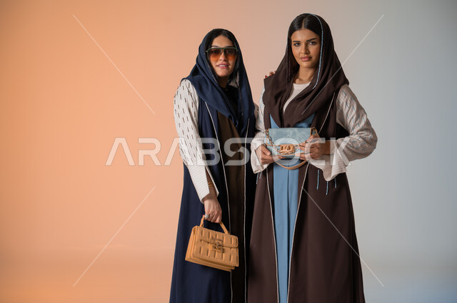اقتناء اكسسوارات ومنتجات لبراندات عالمية، أزياء نسائية عصرية للمحجبات، الاهتمام بالفاشن ومواكبة آخر صيحات الموضة، بورتريه مقرب لصديقتين عربيتين خليجيتين سعوديتين محجبتين ترتديان العباءات الملونة وتمسكان الحقائب الجلدية الفاخرة، وضع النظارات الشمسية، خلفية ملونة