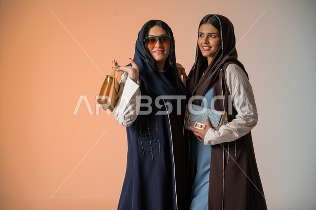 اقتناء اكسسوارات ومنتجات لبراندات عالمية، أزياء نسائية عصرية للمحجبات، الاهتمام بالفاشن ومواكبة آخر صيحات الموضة، بورتريه مقرب لصديقتين عربيتين خليجيتين سعوديتين محجبتين ترتديان العباءات الملونة وتمسكان الحقائب الجلدية الفاخرة، وضع النظارات الشمسية، خلفية ملونة