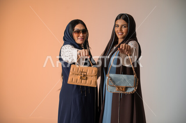 اقتناء اكسسوارات ومنتجات لبراندات عالمية، أزياء نسائية عصرية للمحجبات، الاهتمام بالفاشن ومواكبة آخر صيحات الموضة، بورتريه مقرب لصديقتين عربيتين خليجيتين سعوديتين محجبتين ترتديان العباءات الملونة وتمسكان الحقائب الجلدية الفاخرة، وضع النظارات الشمسية، خلفية ملونة