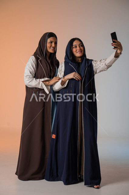 التقاط صور السيلفي رفقة الأصدقاء وقضاء أوقات ممتعة، بورتريه لشابتين عربيتين خليجيتين سعوديتين محجبتين ترتديان العباءات الملونة تقفان وتأخذان اللقطات باستخدام الهاتف المحمول وصنع ذكريات جميلة، الاهتمام بالأناقة والمظهر الخارجي، الجسم بالطول الكامل، خلفية ملونة