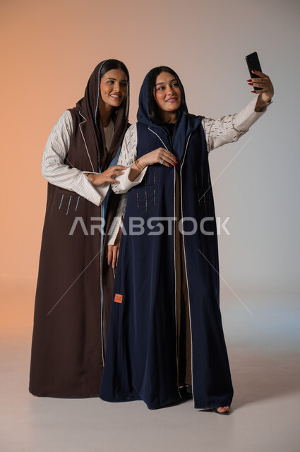 التقاط صور السيلفي رفقة الأصدقاء وقضاء أوقات ممتعة، بورتريه لشابتين عربيتين خليجيتين سعوديتين محجبتين ترتديان العباءات الملونة تقفان وتأخذان اللقطات باستخدام الهاتف المحمول وصنع ذكريات جميلة، الاهتمام بالأناقة والمظهر الخارجي، الجسم بالطول الكامل، خلفية ملونة