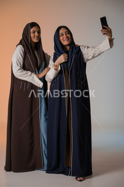 التقاط صور السيلفي رفقة الأصدقاء وقضاء أوقات ممتعة، بورتريه لشابتين عربيتين خليجيتين سعوديتين محجبتين ترتديان العباءات الملونة تقفان وتأخذان اللقطات باستخدام الهاتف المحمول وصنع ذكريات جميلة، الاهتمام بالأناقة والمظهر الخارجي، الجسم بالطول الكامل، خلفية ملونة
