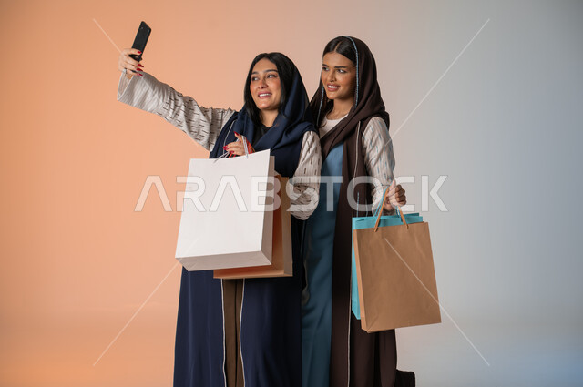 قضاء أوقات ممتعة في التسوق رفقة الأصدقاء، التقاط صور السيلفي بإيماءات السرور والمحبة، بورتريه مقرب لشابتين عربيتين خليجيتين سعوديتين محجبتين ترتديان العباءات الخليجية تمسكان بالأكياس الورقية وتأخذان اللقطات الجميلة باستخدام الهاتف المحمول، خلفية ملونة