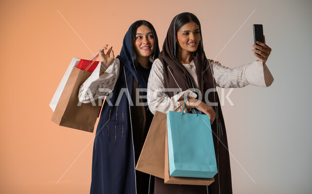 قضاء أوقات ممتعة في التسوق رفقة الأصدقاء، التقاط صور السيلفي بإيماءات السرور والمحبة، بورتريه مقرب لشابتين عربيتين خليجيتين سعوديتين محجبتين ترتديان العباءات الخليجية تمسكان بالأكياس الورقية وتأخذان اللقطات الجميلة باستخدام الهاتف المحمول، خلفية ملونة