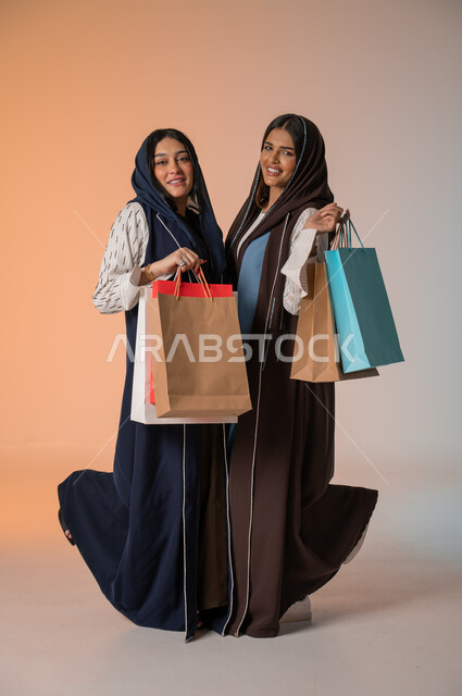 النظر إلى الكاميرا بإيماءات السعادة والسرور، قضاء أوقات ممتعة في التسوق رفقة الأصدقاء، بورتريه لشابتين عربيتين خليجيتين سعوديتين محجبتين ترتديان العباءات الخليجية تحملان العديد من الأكياس الورقية، شراء المستلزمات والهدايا، الجسم بالطول الكامل، خلفية ملونة